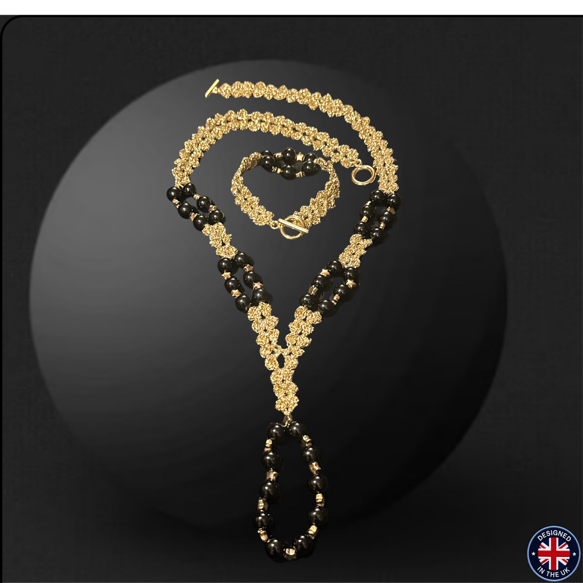 A gold and black beaded necklace displayed on a dark studio background, with a matching bracelet also visible. The necklace features textured gold floral-like links mixed with strands of glossy black beads, arranged in a draped display.