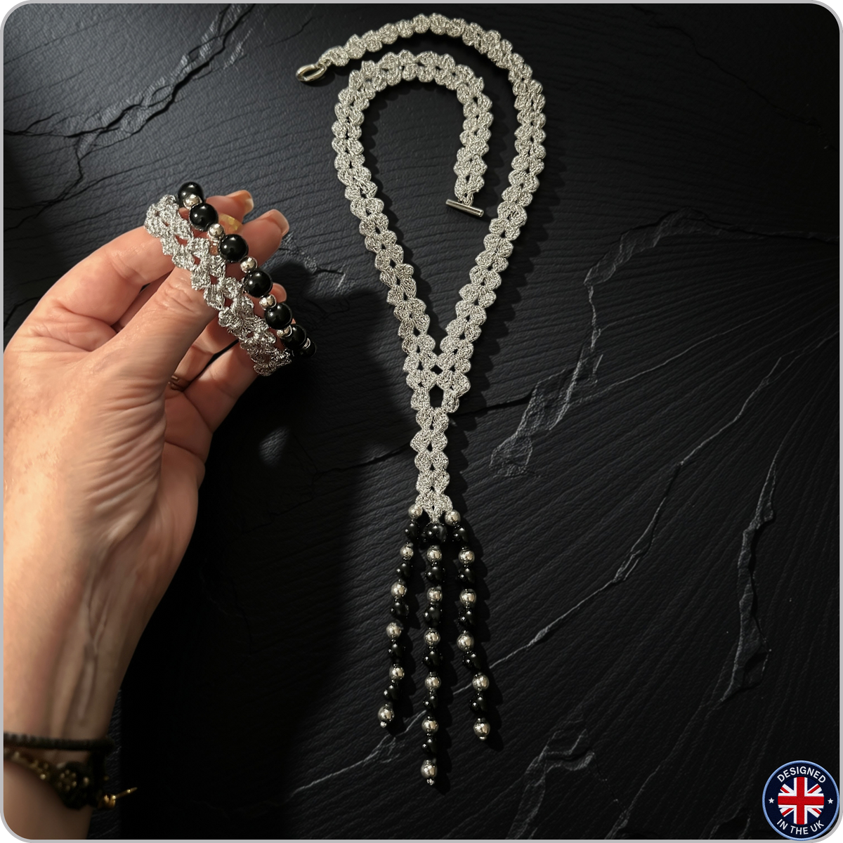 A handmade silver and black beaded necklace with a matching multi-strand bracelet is displayed on a dark slate background. A hand holds the bracelet on the left, and a small UK-made badge is in the bottom right corner.