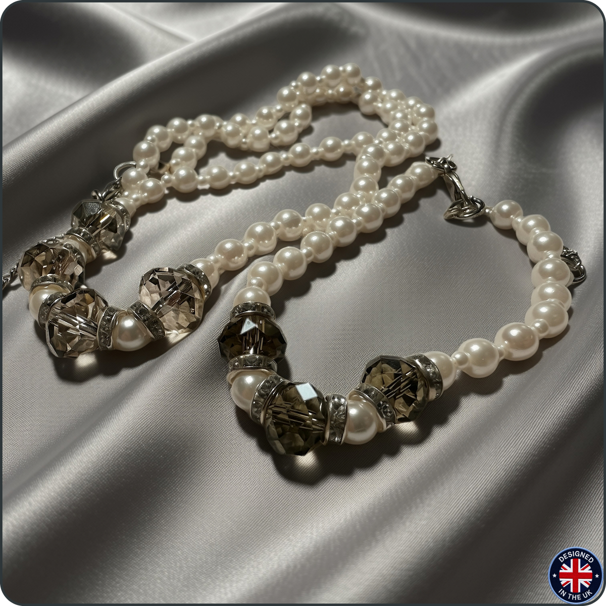 A pearl and crystal beaded necklace with silver-toned spacers, arranged on draped satin fabric. The necklace features white pearl beads alternating with faceted smoky crystal beads and silver-toned metal spacers.