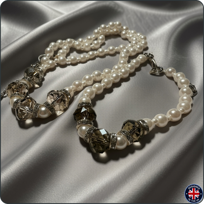 A pearl and crystal beaded necklace with silver-toned spacers, arranged on draped satin fabric. The necklace features white pearl beads alternating with faceted smoky crystal beads and silver-toned metal spacers.