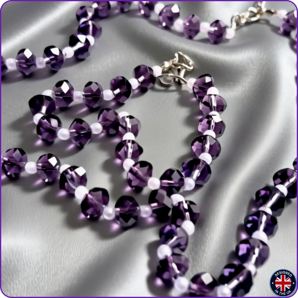 A beaded necklace with faceted purple beads and small white spacer beads, draped on a shiny silver satin fabric background. A silver clasp is visible at the top center.