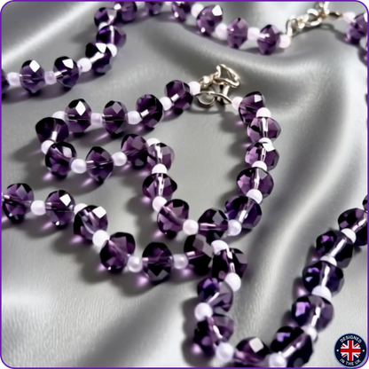 A beaded necklace with faceted purple beads and small white spacer beads, draped on a shiny silver satin fabric background. A silver clasp is visible at the top center.