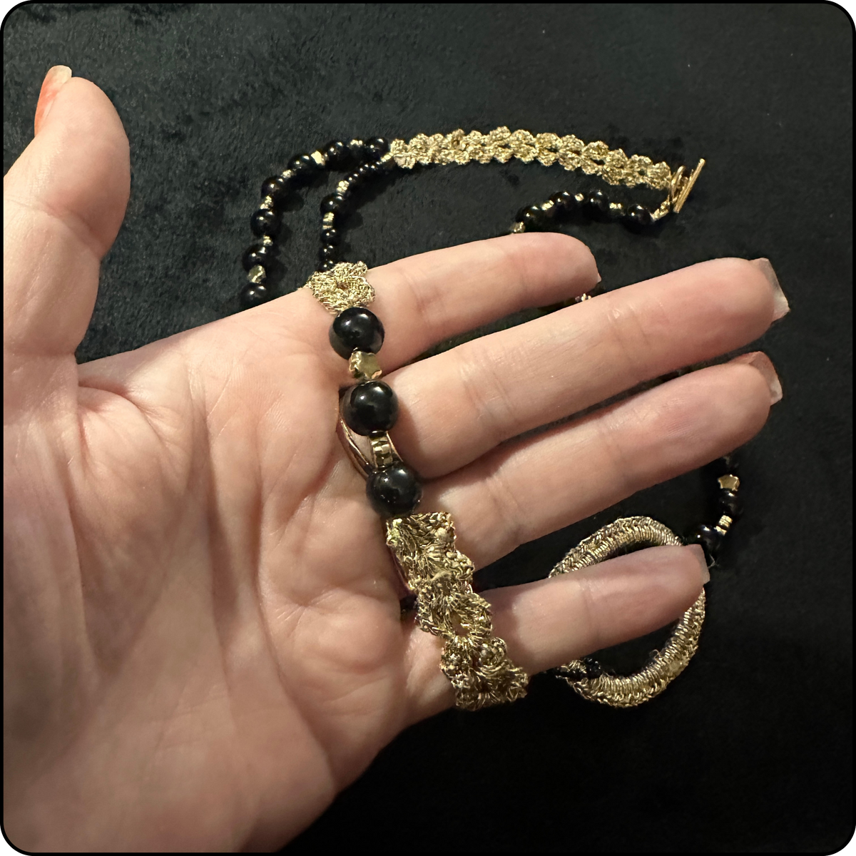 A gold and black beaded necklace with textured gold elements, held in a person's hand against a black background.
