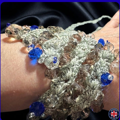 A close-up of a handmade beaded bracelet worn on a wrist. The bracelet features textured, knotted light grey crochet with faceted clear beads and bright blue beads.
