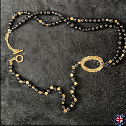 A double-strand beaded necklace featuring black faceted beads, small gold star-shaped beads, and gold-toned metal accents, including a large oval centerpiece and a toggle clasp, displayed on a dark textured background.