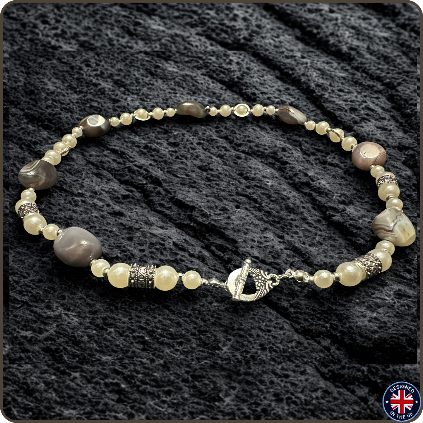 A beaded necklace with a mix of cream, gray, and brown beads, accented by textured silver spacers and a silver toggle clasp, displayed on a dark textured fabric background.