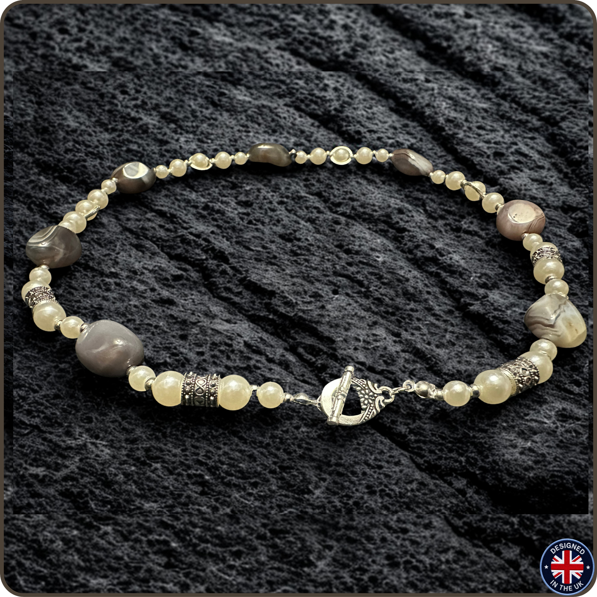 A beaded necklace with a mix of cream, gray, and brown beads, accented by textured silver spacers and a silver toggle clasp, displayed on a dark textured fabric background.