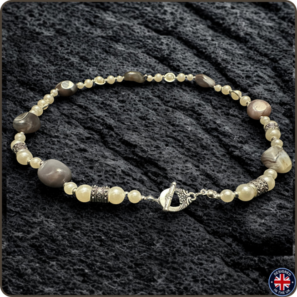 A beaded necklace with a mix of cream, gray, and brown beads, accented by textured silver spacers and a silver toggle clasp, displayed on a dark textured fabric background.
