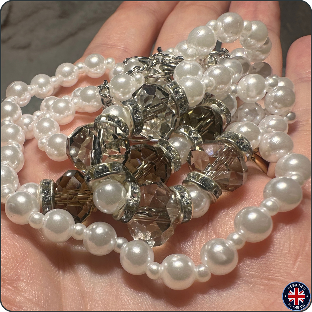 A close-up of a hand holding a strand of white faux-pearl beads with faceted clear crystal spacers and silver-toned metal rings. A small circular 'Made in UK' badge is visible in the bottom right corner.