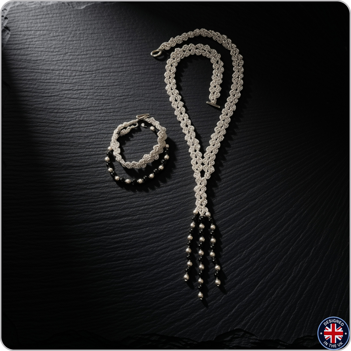 A silver-toned beaded jewelry set consisting of a long beaded necklace and a matching beaded bracelet, displayed on a dark textured slate background.