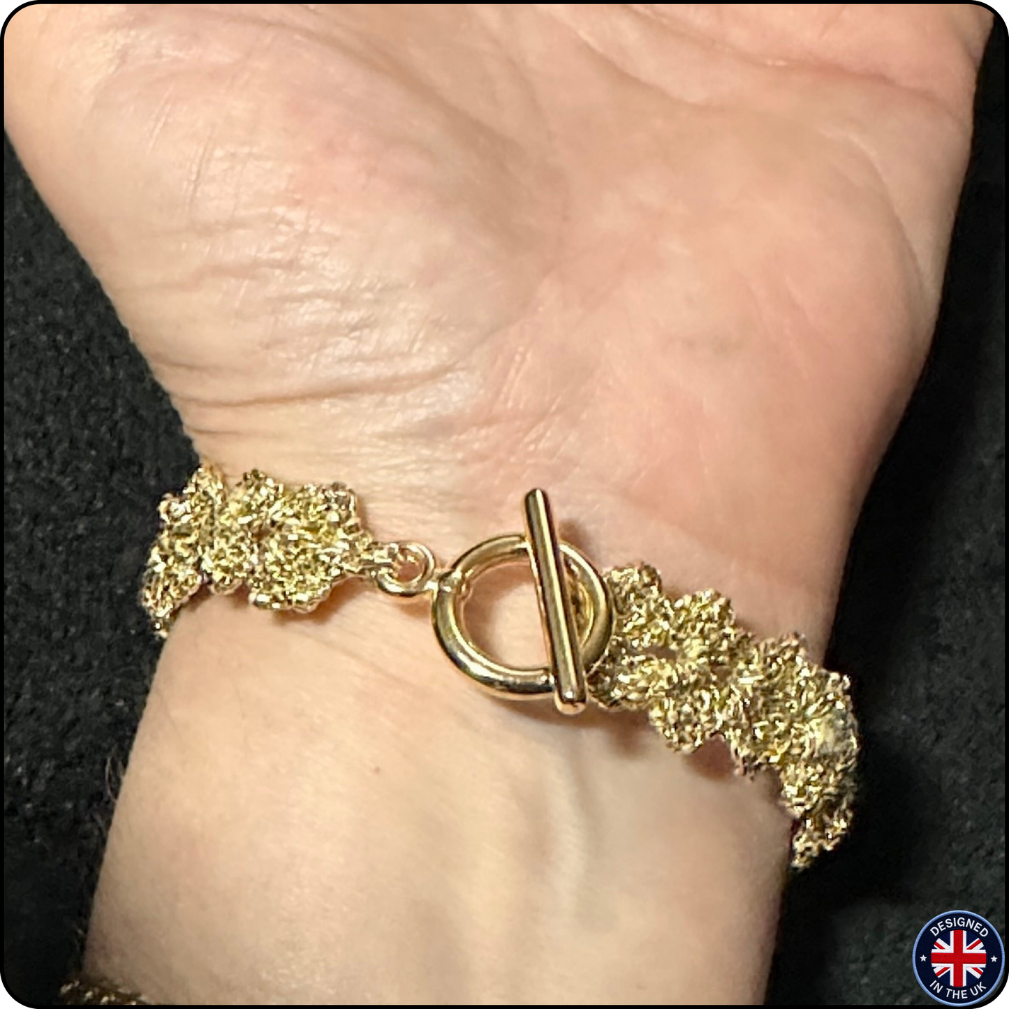 A close-up of a gold-toned textured bracelet with a toggle clasp worn on a person's wrist against a dark background.