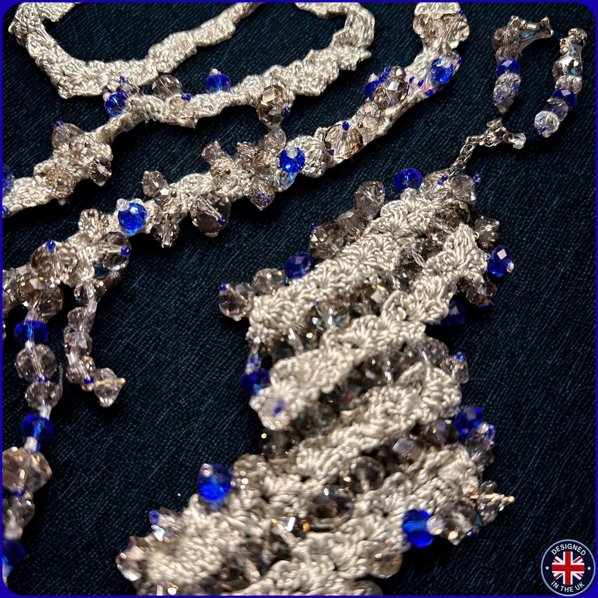 A close-up of a beaded tassel necklace with intricate silver crochet or beadwork, clear faceted beads, and bright blue rhinestone accents.