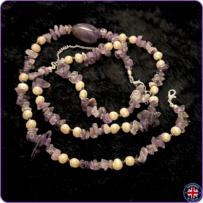 A beaded necklace featuring light cream-colored round pearls and lavender-purple translucent stone chips, with a polished oval purple bead and silver-tone clasp, displayed on a black textured background.