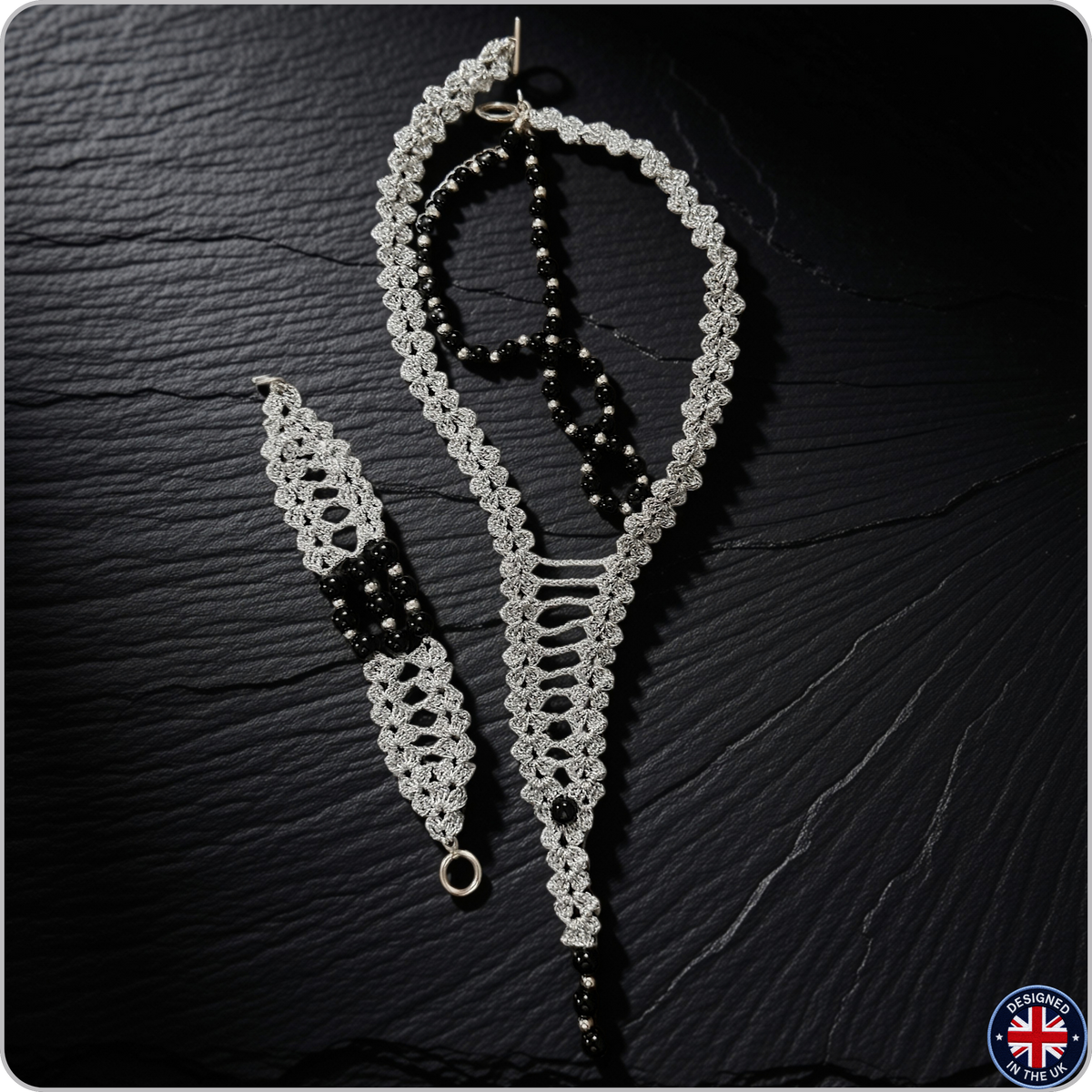 A handmade macrame jewelry set featuring a long necklace, a matching bracelet, and a dangling earring, crafted with white and black macrame cords and black beads, displayed on a dark textured slate background.