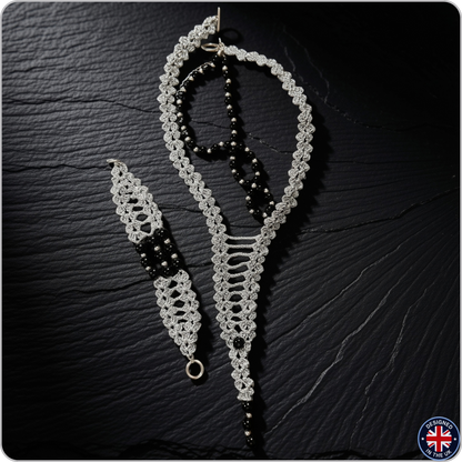 A handmade macrame jewelry set featuring a long necklace, a matching bracelet, and a dangling earring, crafted with white and black macrame cords and black beads, displayed on a dark textured slate background.