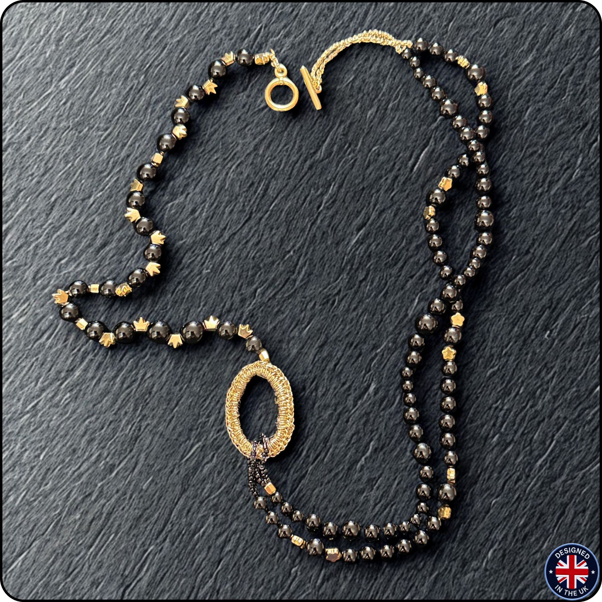 A double-strand beaded necklace featuring faceted black and metallic gold beads, with a textured gold oval pendant and a gold clasp, displayed on a dark slate background.