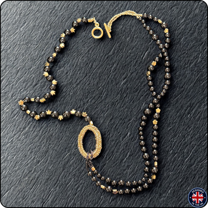A double-strand beaded necklace featuring faceted black and metallic gold beads, with a textured gold oval pendant and a gold clasp, displayed on a dark slate background.