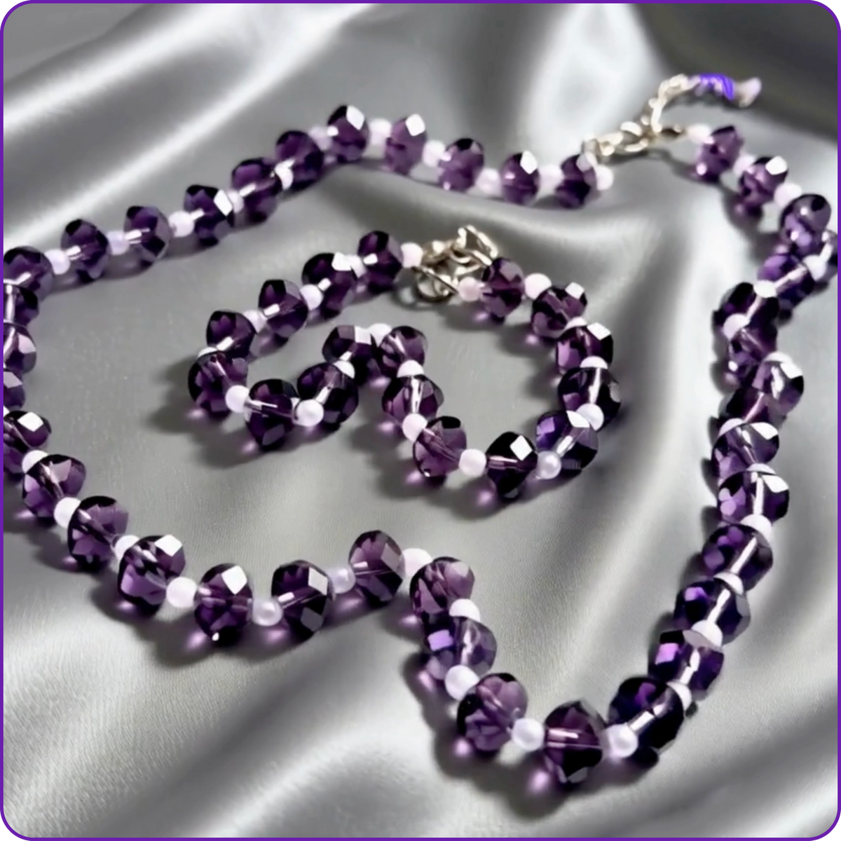 A matching beaded necklace and bracelet set in faceted purple beads with small white accent beads, displayed on a draped silver satin fabric background.