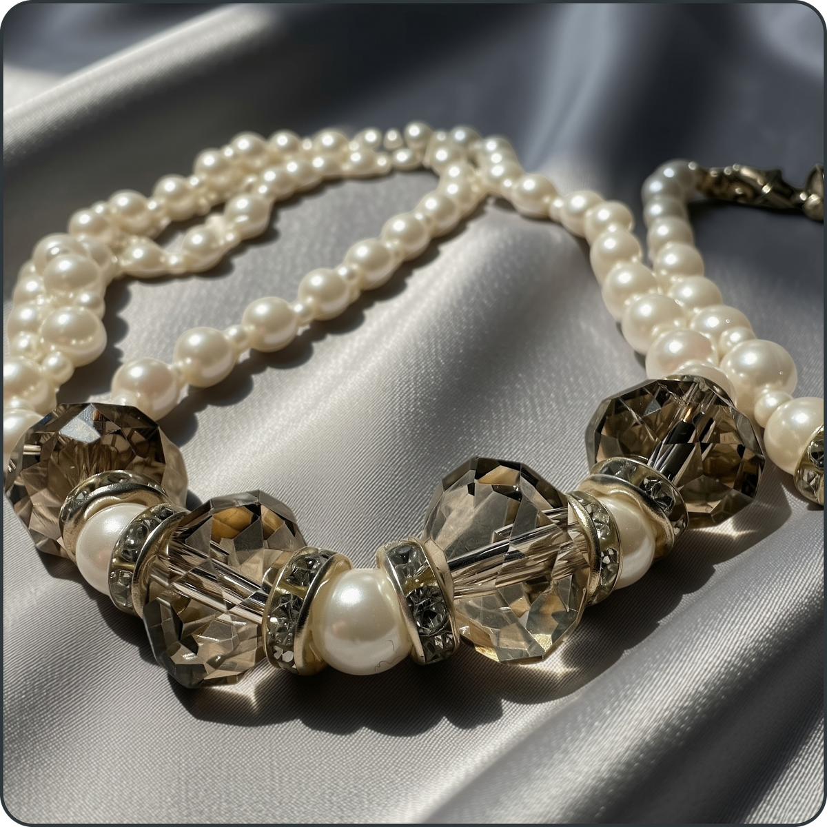 A close-up of a pearl and crystal statement necklace draped on draped satin fabric. The necklace features multiple strands of creamy white faux pearls and large faceted champagne-colored crystal beads with gold-toned spacers.