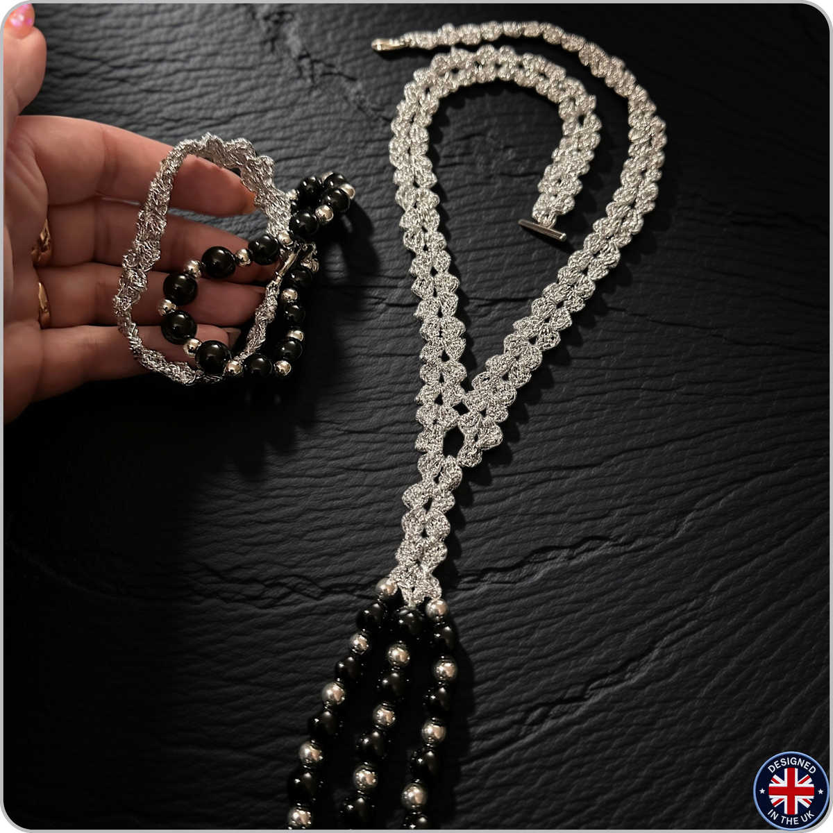 A silver beaded necklace and matching bracelet are displayed on a dark textured surface, with a hand holding the bracelet to show its scale. The jewelry features intricate silver beadwork and glossy black beads.