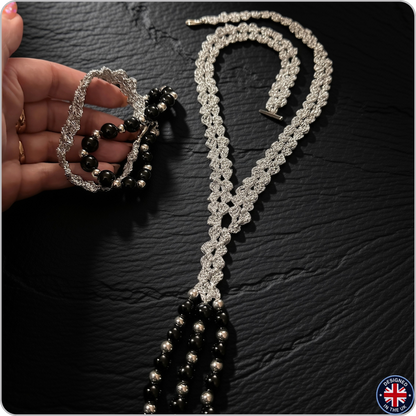 A silver beaded necklace and matching bracelet are displayed on a dark textured surface, with a hand holding the bracelet to show its scale. The jewelry features intricate silver beadwork and glossy black beads.