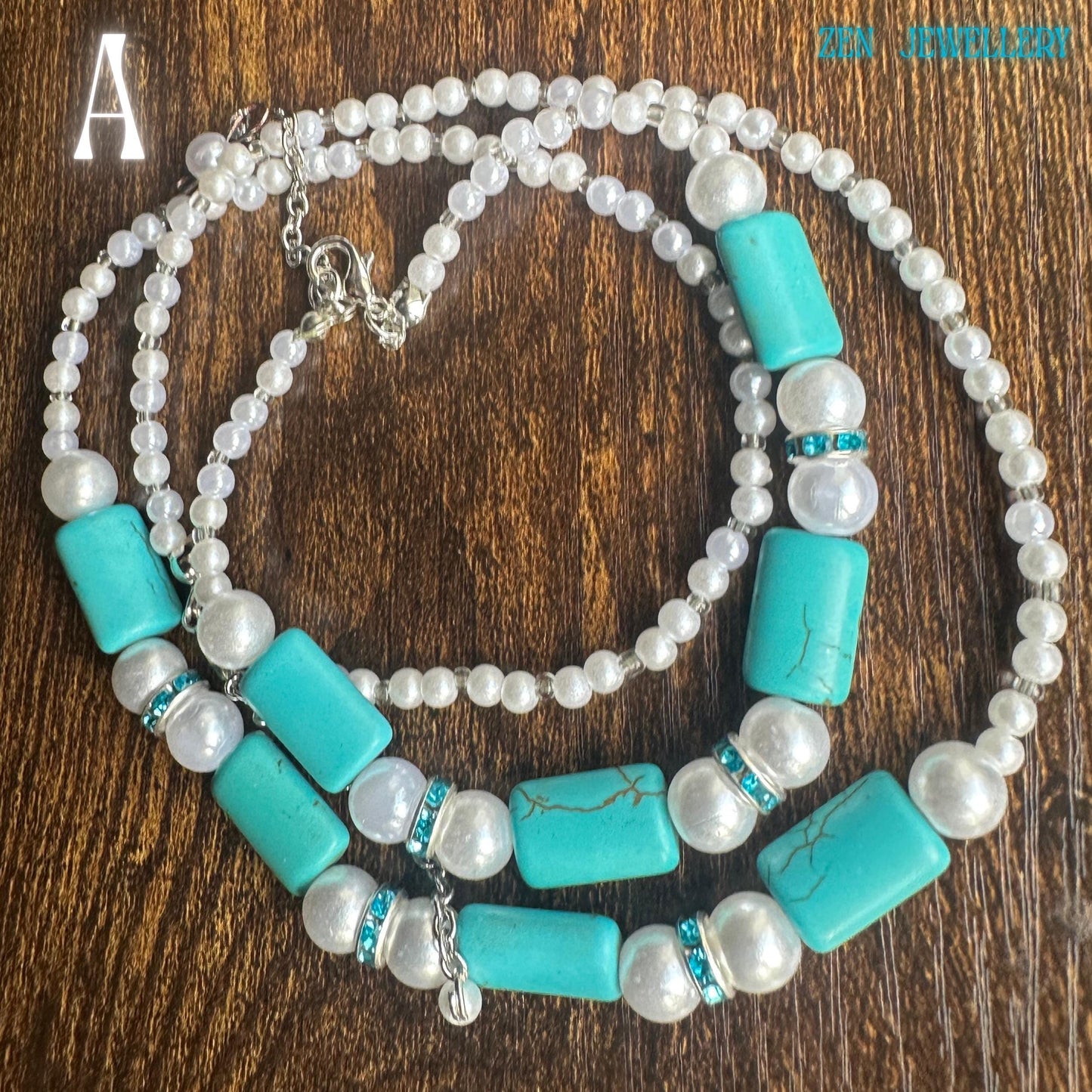 A layered necklace with turquoise rectangular beads, white faux pearls, and small white beads. The necklace has silver-toned clasps and accents, with a small charm. The turquoise beads have a natural, cracked appearance. The necklace is displayed on a brown surface.