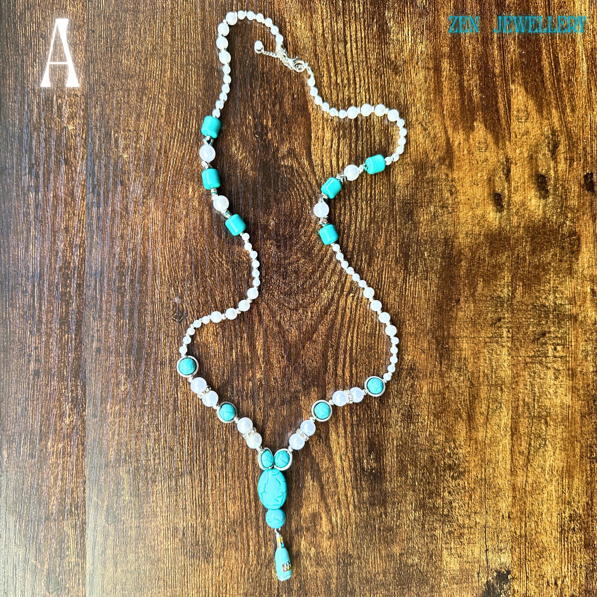 The handcrafted TURQUOISE PEARL necklace is photographed diagonally to emphasise its flowing design and balanced arrangement of turquoise pebbles, pearls, and sparkling silver accents. The rich, earthy wooden background enhances the vivid blue-green stones while the elegant tassel pendant creates movement. The necklace measures approximately 64 cm long, making it ideal for layering or standalone statement styling.