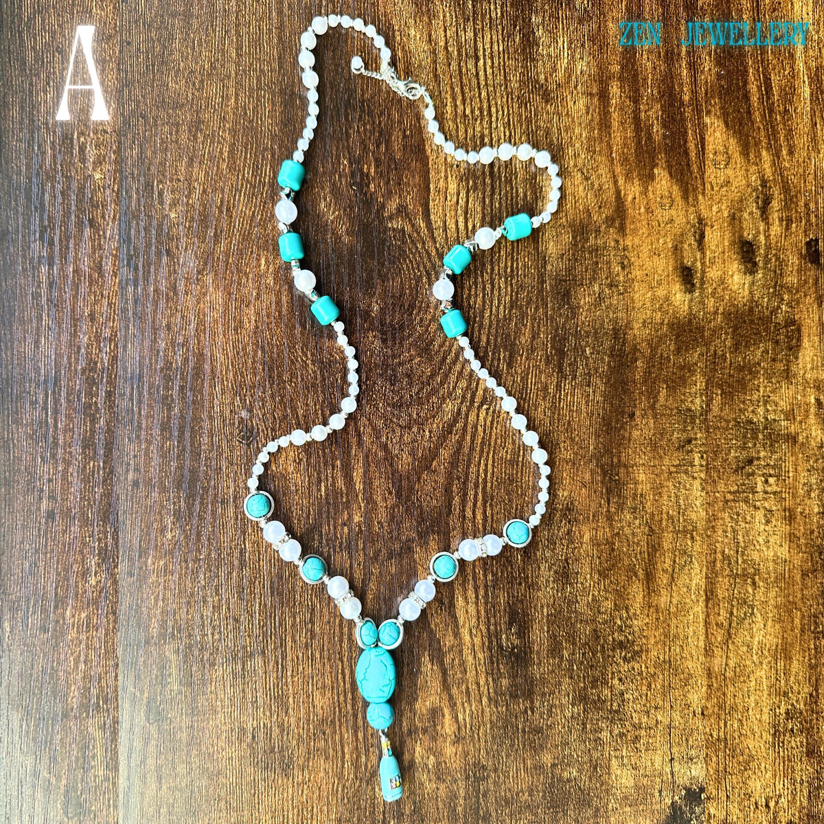 The handcrafted TURQUOISE PEARL necklace is photographed diagonally to emphasise its flowing design and balanced arrangement of turquoise pebbles, pearls, and sparkling silver accents. The rich, earthy wooden background enhances the vivid blue-green stones while the elegant tassel pendant creates movement. The necklace measures approximately 64 cm long, making it ideal for layering or standalone statement styling.