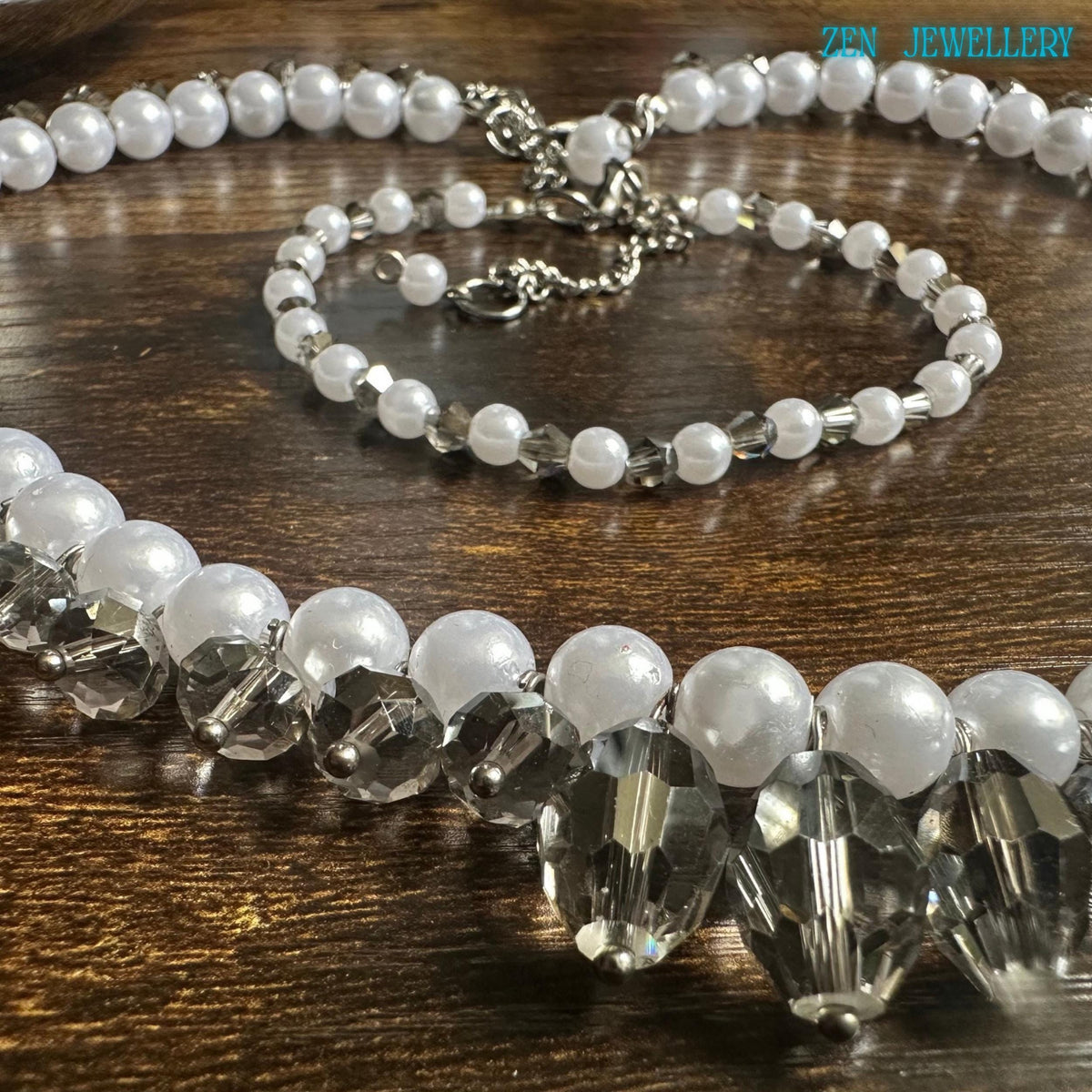 Sophisticated bridal-inspired jewellery set made with white pearl-style beads sparkling crystal accents and silver closures arranged on a dark textured surface for strong visual contrast. The necklace forms a dramatic curved shape with crystal drop details at the centre front while the matching bracelet creates a delicate complementary style. The glossy pearl finish and reflective crystals create an elegant luxurious appearance for weddings eveni