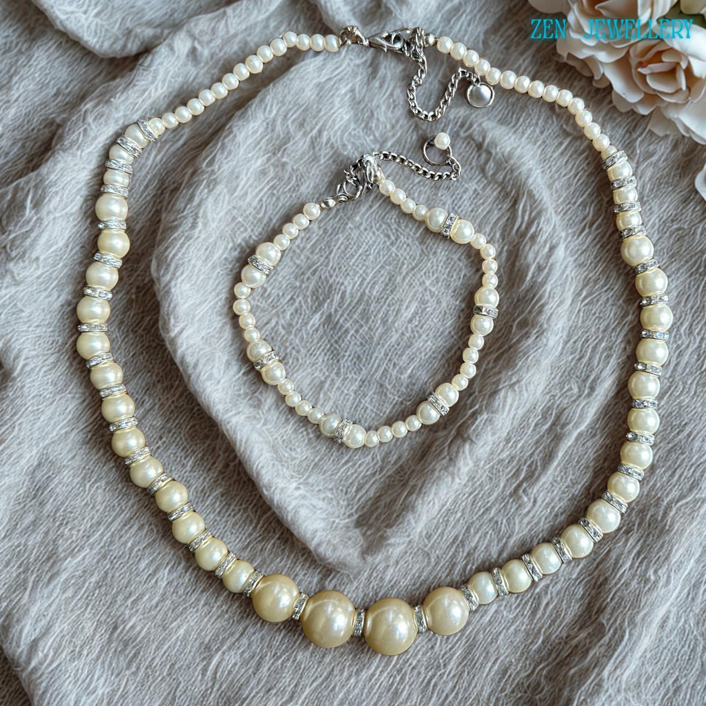 Handmade white pearl jewellery set with glossy ivory beads sparkling silver rhinestone spacers and adjustable silver chain closures arranged on a soft neutral fabric background. The necklace features larger central pearls surrounded by smaller round beads creating balance texture and elegance while the matching bracelet complements the necklace perfectly. The soft pearl finish reflects light beautifully creating a graceful, feminine appearance id