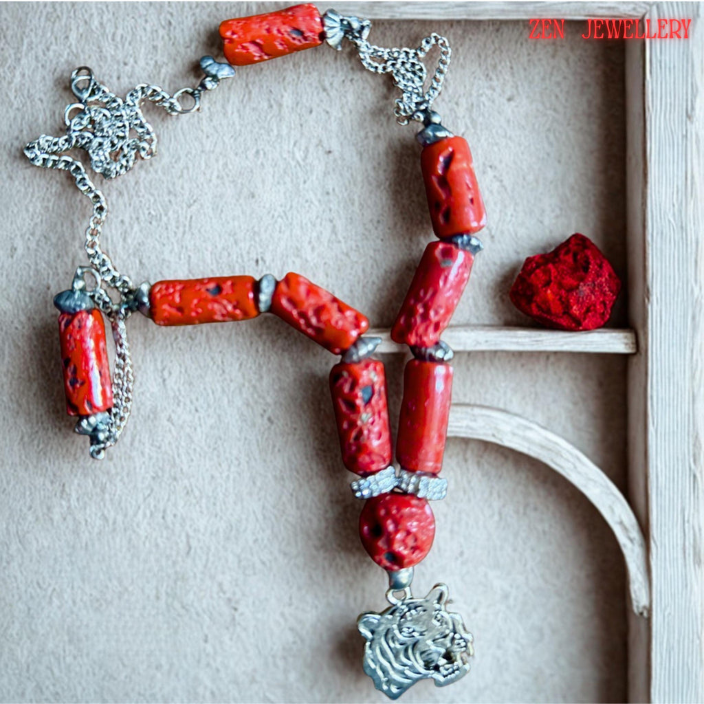 Artistic jewellery photo showing the bold red coral necklace arranged symmetrically against dark wood. The vibrant coral cylinders contrast beautifully with the silver chain and metallic tiger pendant creating a strong bohemian statement piece. The necklace includes textured coral beads, rhinestone separators, and silver accents designed for everyday bold styling or gifting.