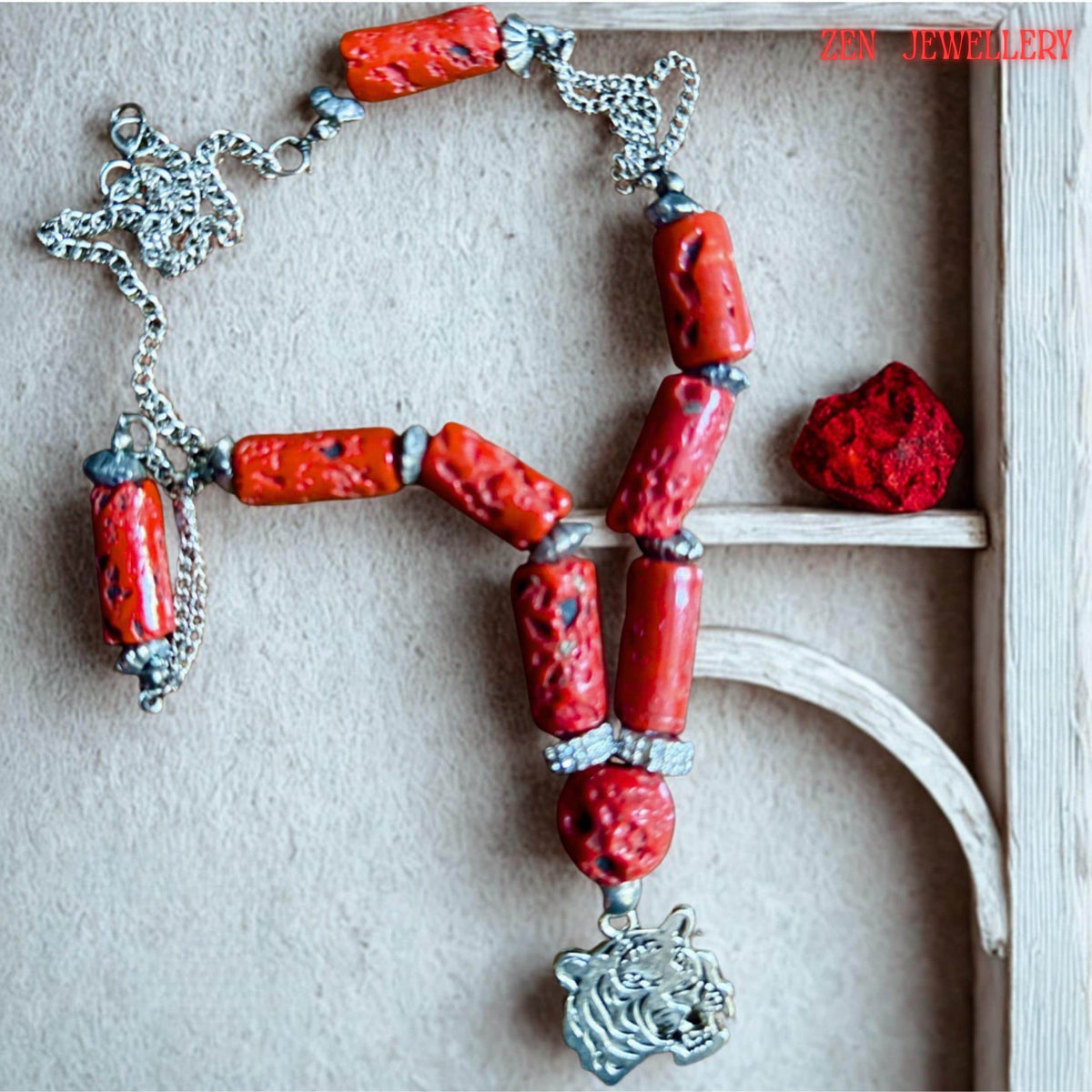 Artistic jewellery photo showing the bold red coral necklace arranged symmetrically against dark wood. The vibrant coral cylinders contrast beautifully with the silver chain and metallic tiger pendant creating a strong bohemian statement piece. The necklace includes textured coral beads, rhinestone separators, and silver accents designed for everyday bold styling or gifting.