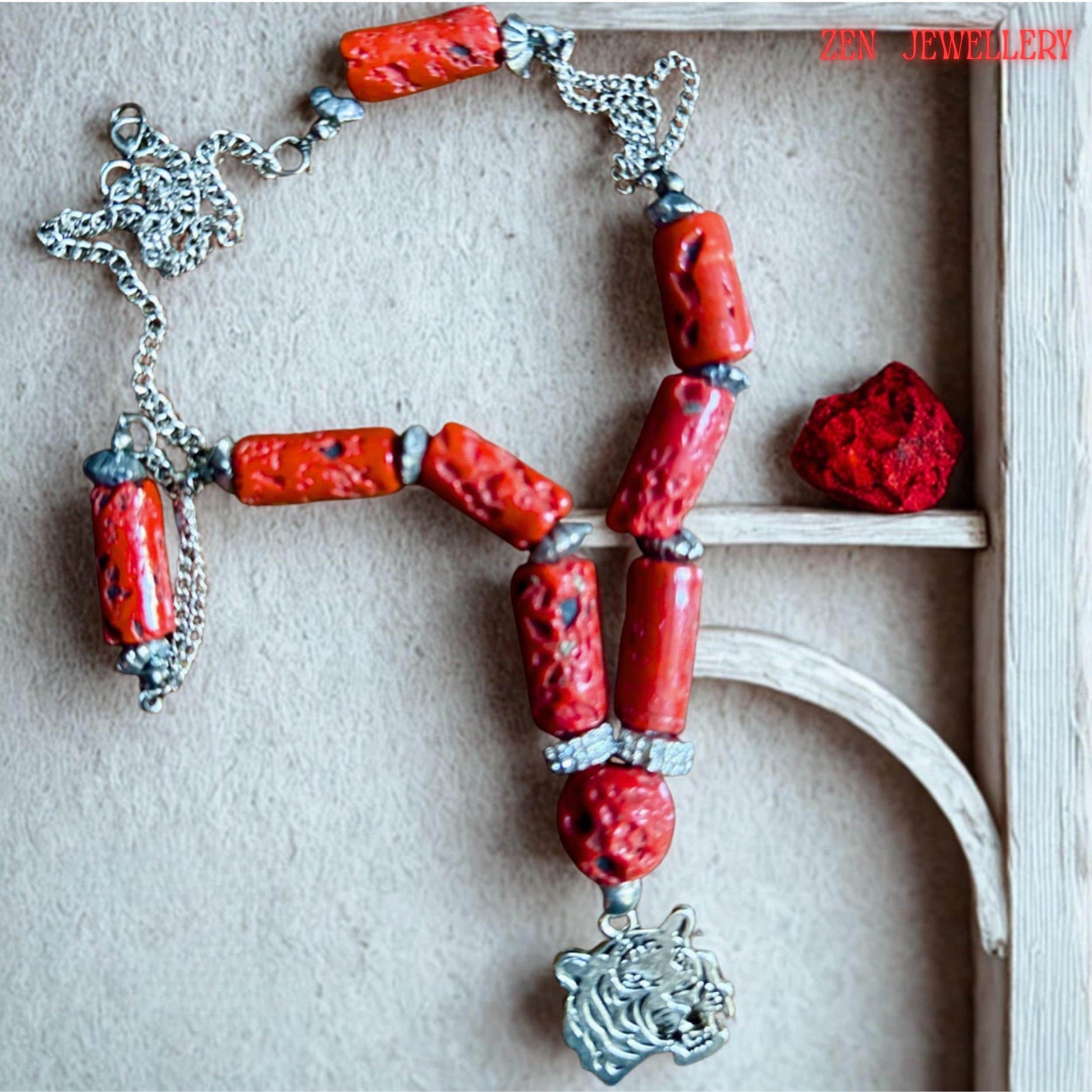 Artistic jewellery photo showing the bold red coral necklace arranged symmetrically against dark wood. The vibrant coral cylinders contrast beautifully with the silver chain and metallic tiger pendant creating a strong bohemian statement piece. The necklace includes textured coral beads, rhinestone separators, and silver accents designed for everyday bold styling or gifting.