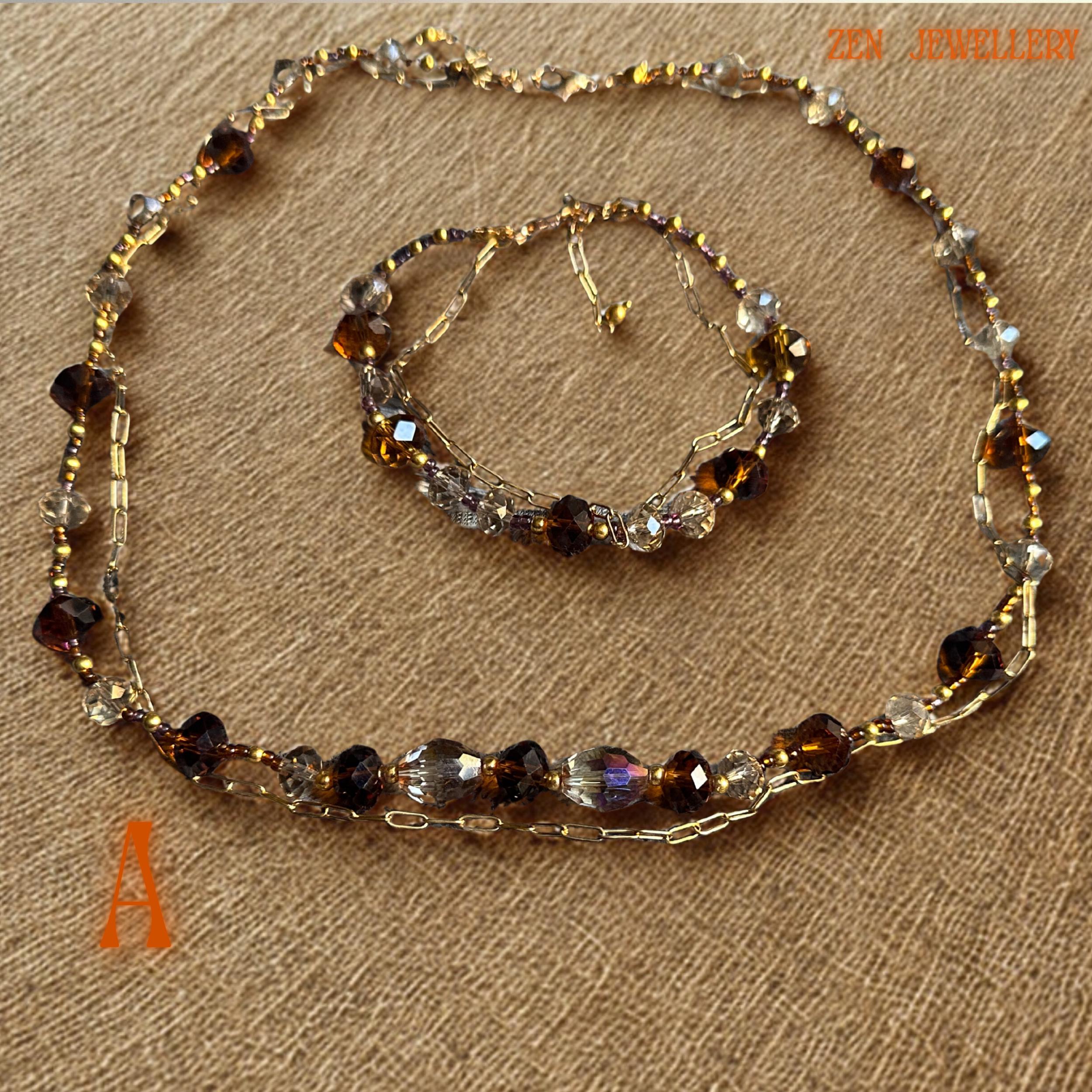 Detailed image showing faceted caramel and bronze Austrian crystal beads with reflective surfaces that shimmer under soft lighting. Gold spacer beads and elegant clasp details create a refined finish while the layered chain sections add texture and artistic movement. The matching bracelet complements the necklace perfectly and creates a coordinated luxurious jewellery look.