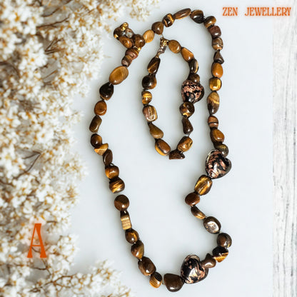 Brown tiger eye necklace and bracelet set displayed on a neutral marble surface with dried floral decoration highlighting the warm amber gemstone colours and delicate gold details. The smooth, polished stones create texture, shine, and a refined handcrafted appearance.
