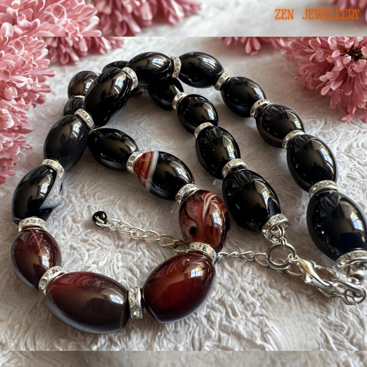 Elegant black and burgundy agate gemstone necklace displayed on a soft cream textured background with pink floral decoration. The smooth oval beads have a glossy reflective finish while the striped reddish-brown agate stones add natural texture and depth. Sparkling rhinestone spacer rings create luxurious detail between each bead. Finished with a silver clasp and extension chain. Necklace length is 47 cm approximately 18.5 inches.