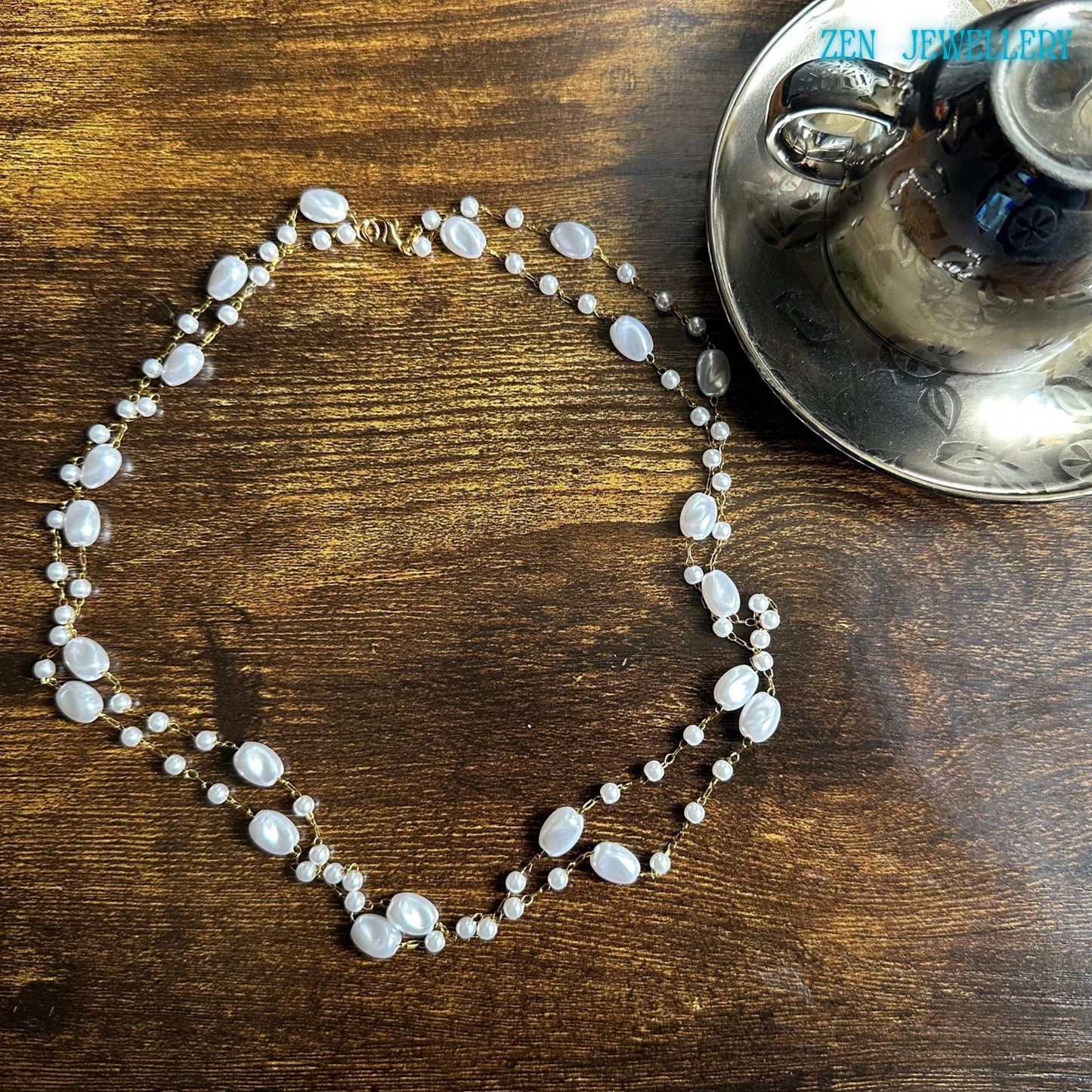 This feminine handmade necklace combines white pearl-style beads in different sizes with elegant golden chain links for a soft luxurious look. The asymmetrical arrangement gives the necklace an organic artistic style inspired by modern boho jewellery and minimalist fashion. The glossy beads create beautiful texture and shine against the dark wooden surface.