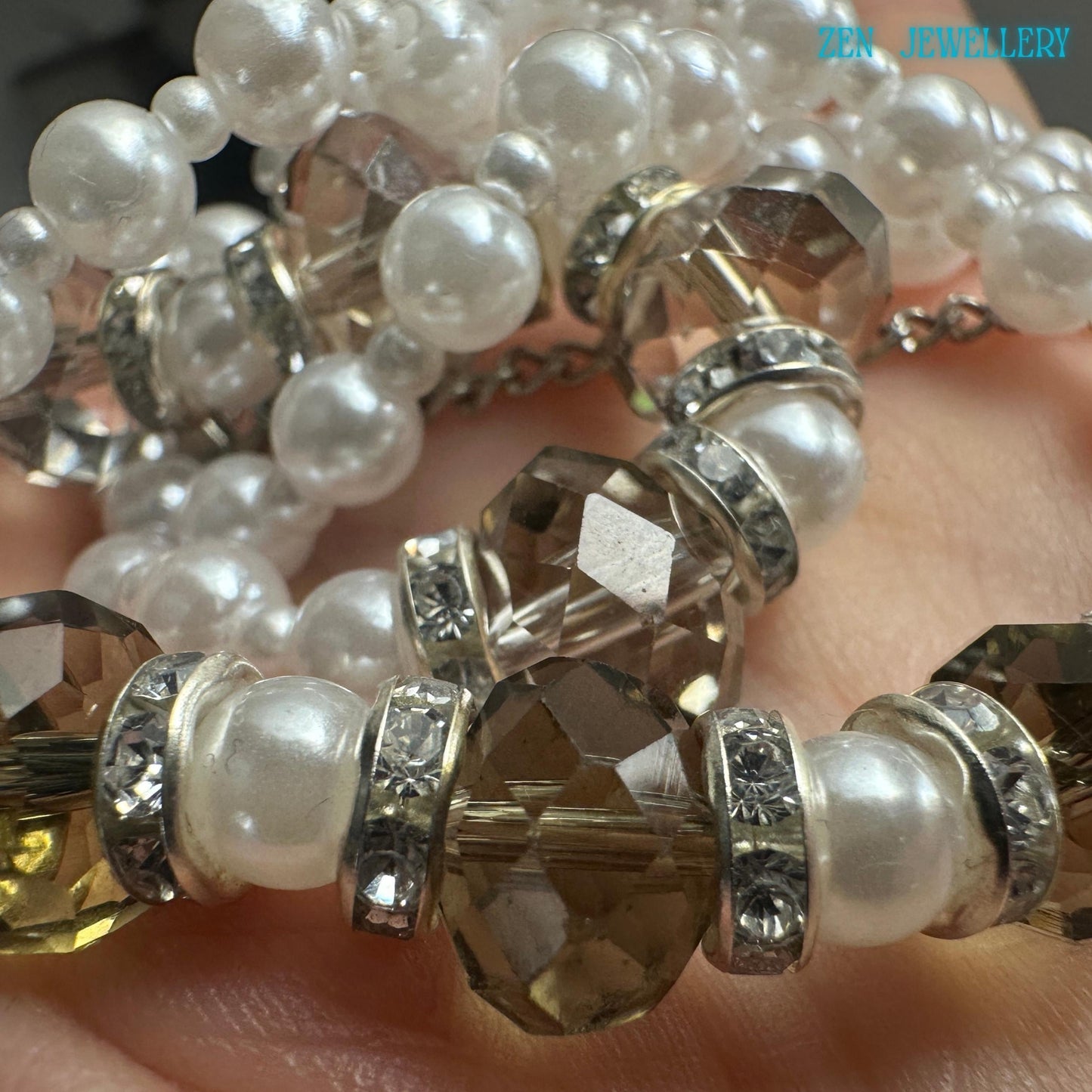 Elegant white pearl necklace and bracelet set photographed flat against a textured dark surface. The necklace features rows of small pearl-style beads combined with larger glossy white accents and faceted smoky crystal beads. Silver rhinestone spacers separate the beads and create a refined sparkling effect. The bracelet mirrors the necklace design for a coordinated and polished jewellery look.