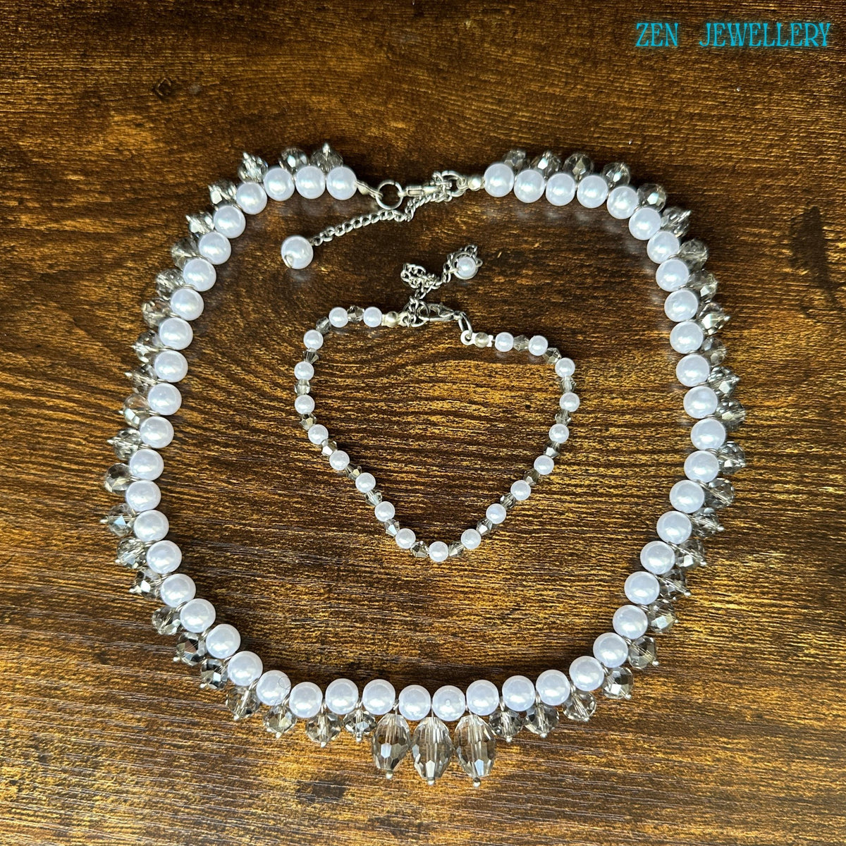 Handmade bridal jewellery set featuring white pearl-style beads paired with shimmering faceted crystals and elegant silver spacer accents. The necklace creates a bold, curved silhouette with dangling crystal details, while the bracelet adds a coordinated, delicate touch. The glossy pearls contrast beautifully against the dark background, and the silver chain closures provide adjustable comfort.