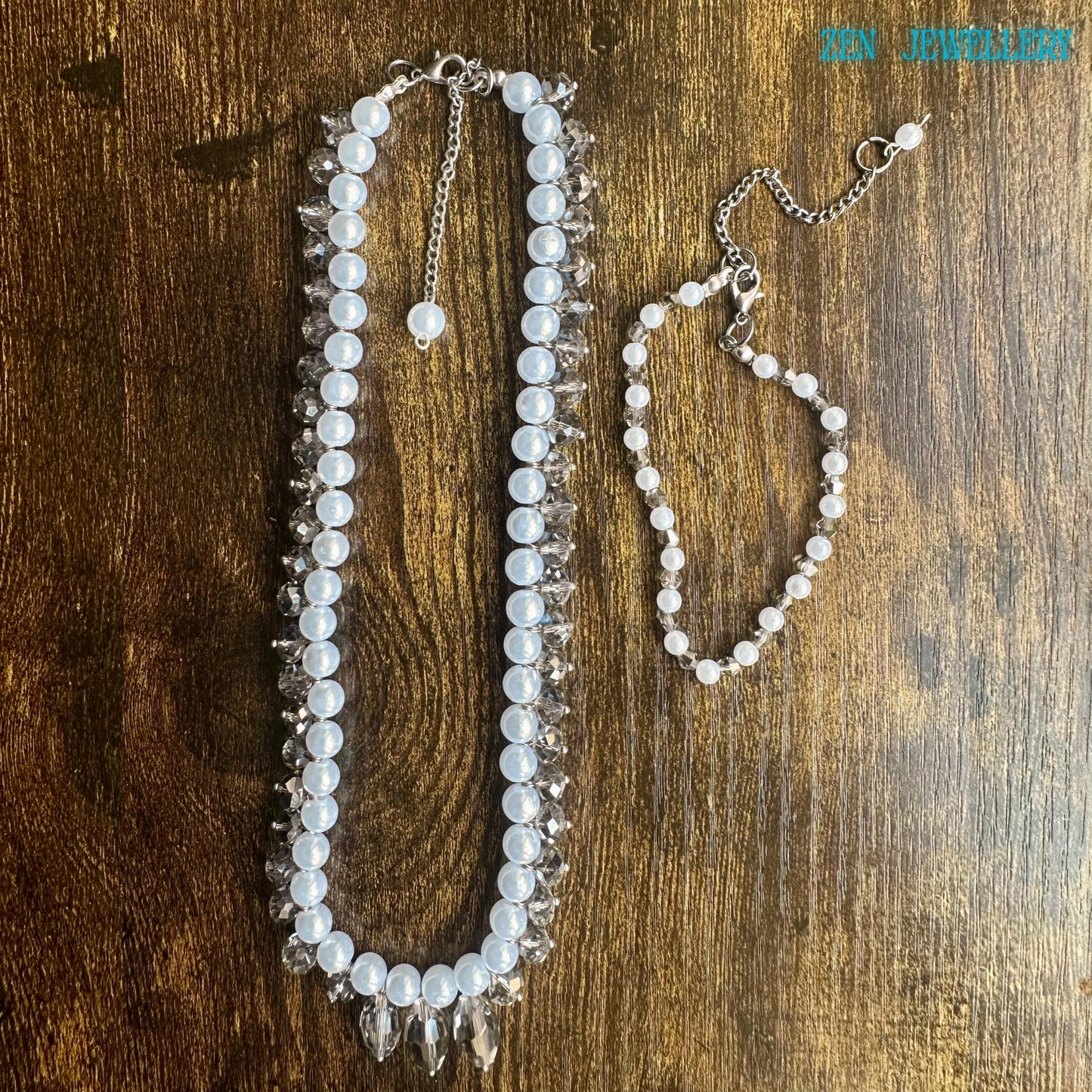 White pearl jewellery set with faceted crystal accents and silver chain clasp displayed in an artistic circular arrangement on a dark textured background. The handmade necklace features luminous pearl-style beads and sparkling champagne-grey crystals that reflect light from different angles creating a glamorous yet timeless look. The matching bracelet adds elegance and balance making the set ideal for bridal wear romantic fashion and special occa