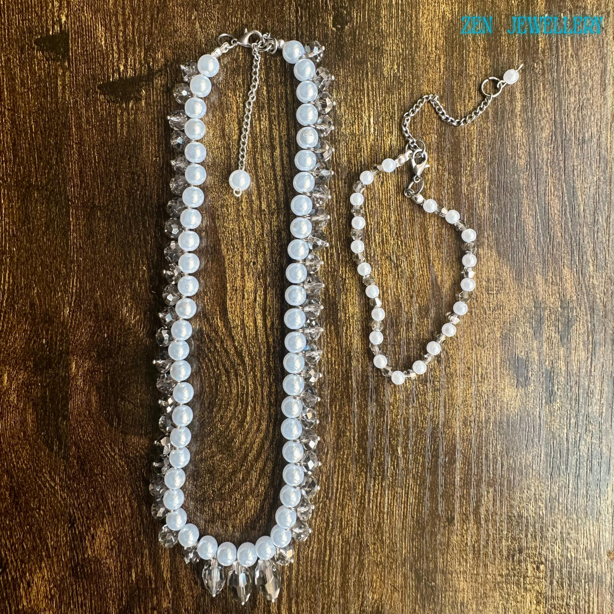 White pearl jewellery set with faceted crystal accents and silver chain clasp displayed in an artistic circular arrangement on a dark textured background. The handmade necklace features luminous pearl-style beads and sparkling champagne-grey crystals that reflect light from different angles creating a glamorous yet timeless look. The matching bracelet adds elegance and balance making the set ideal for bridal wear romantic fashion and special occa
