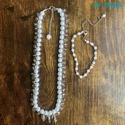White pearl jewellery set with faceted crystal accents and silver chain clasp displayed in an artistic circular arrangement on a dark textured background. The handmade necklace features luminous pearl-style beads and sparkling champagne-grey crystals that reflect light from different angles creating a glamorous yet timeless look. The matching bracelet adds elegance and balance making the set ideal for bridal wear romantic fashion and special occa