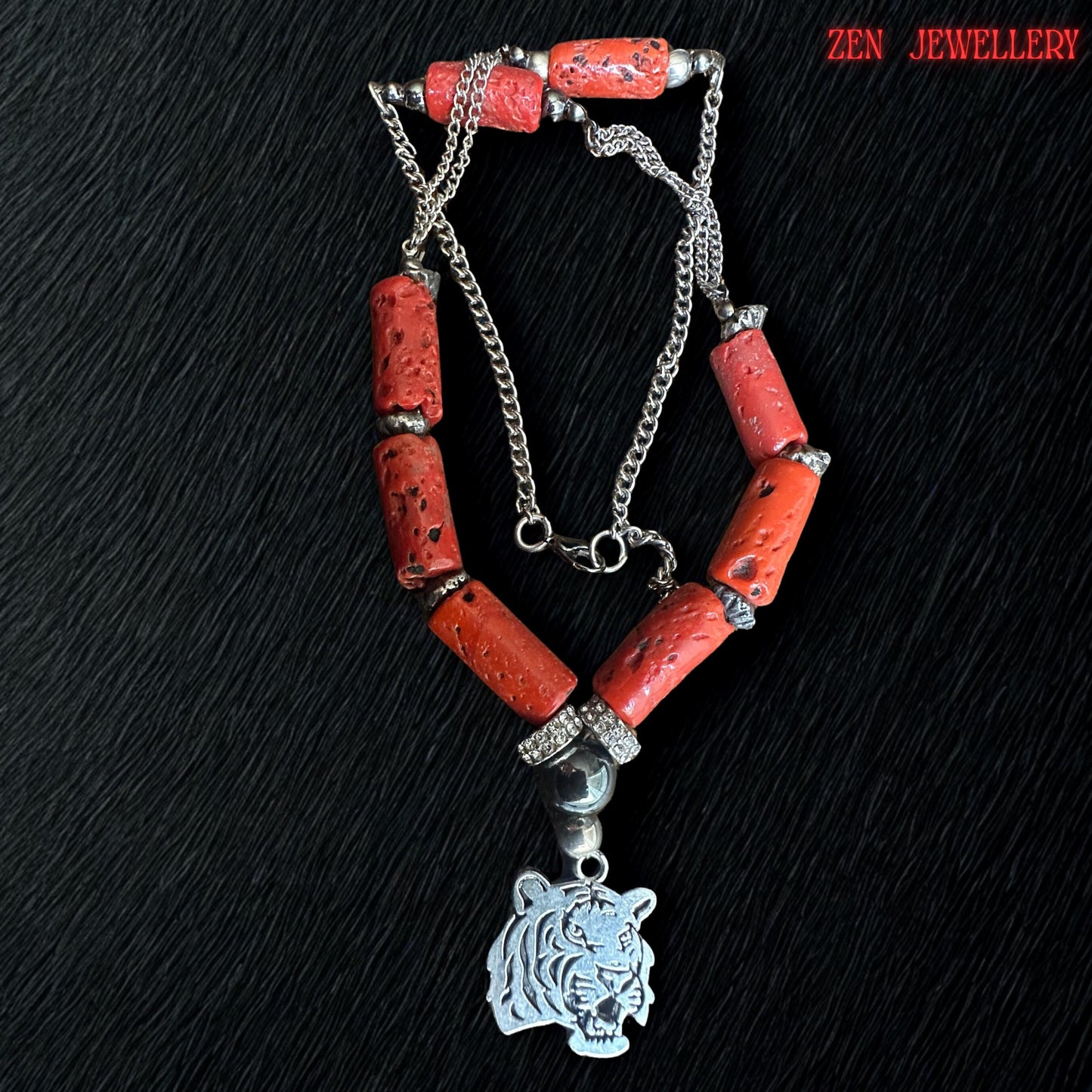Detailed image highlighting the silver tiger pendant attached to the red coral necklace. The pendant features engraved tiger facial details with polished silver finish beneath black glossy beads and sparkling rhinestone spacers. The surrounding red coral beads show raw organic texture and natural imperfections that enhance the handmade artisan appearance. Necklace length is approximately 22.8 inches.