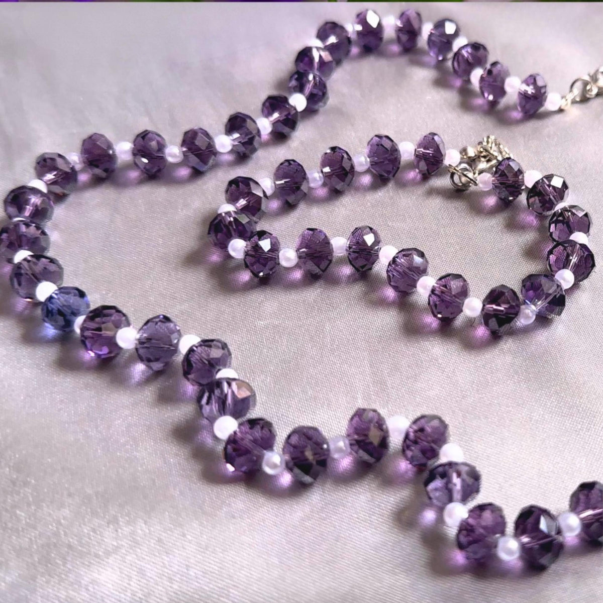 Romantic handmade crystal necklace set featuring rich amethyst purple glass beads combined with small white pearl spacer beads and silver clasp details arranged on soft neutral satin fabric for bridal inspired styling.