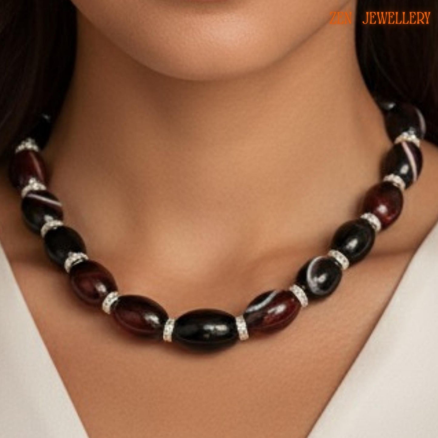 Detailed image of AGATE GEMSTONE necklace showing polished oval black beads with reflective shine and warm burgundy striped agate stones. Rhinestone spacer rings create sparkling silver accents between each gemstone bead. The adjustable silver extension chain adds flexibility and elegant finishing detail. Displayed on a textured fabric surface with soft pink flowers. Necklace size is approximately 47 cm or 18.5 inches long.
