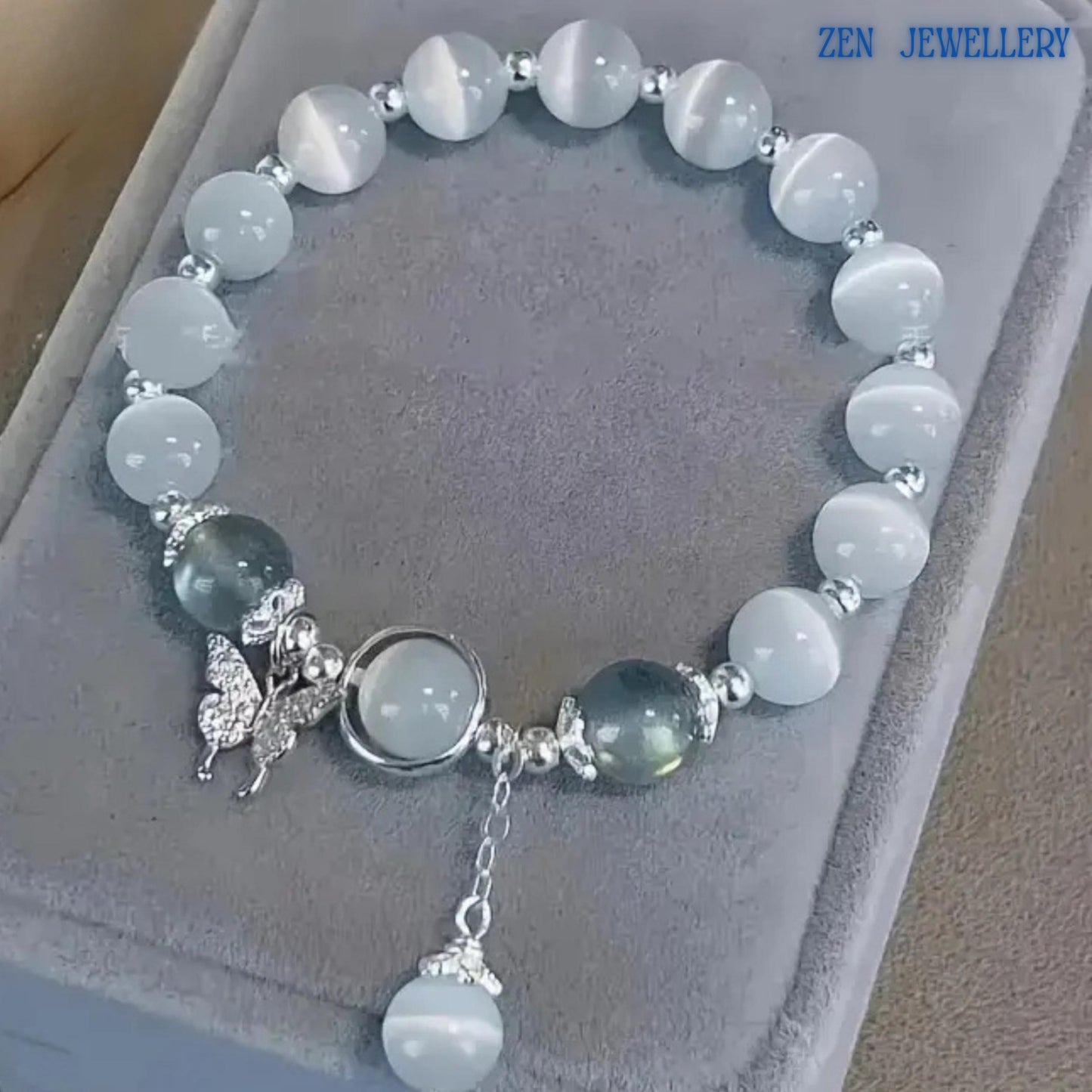 Bracelet photographed from above showing the complete circular arrangement of white gemstone beads with soft glowing appearance, grey gemstone accents placed strategically for contrast, silver decorative elements and a dangling gemstone charm suspended from a delicate chain, allowing buyers to appreciate the overall composition, proportions and elegant styling of the handcrafted jewellery piece.