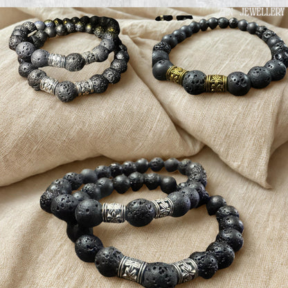 Minimalist black lava stone jewellery set photographed on a textured beige background to emphasise the deep black volcanic beads and polished silver spacer details. The bracelet features natural porous stones with organic matte surfaces and a handcrafted arrangement. The combination of volcanic texture and silver accents creates a stylish balance between earthy simplicity and refined elegance.