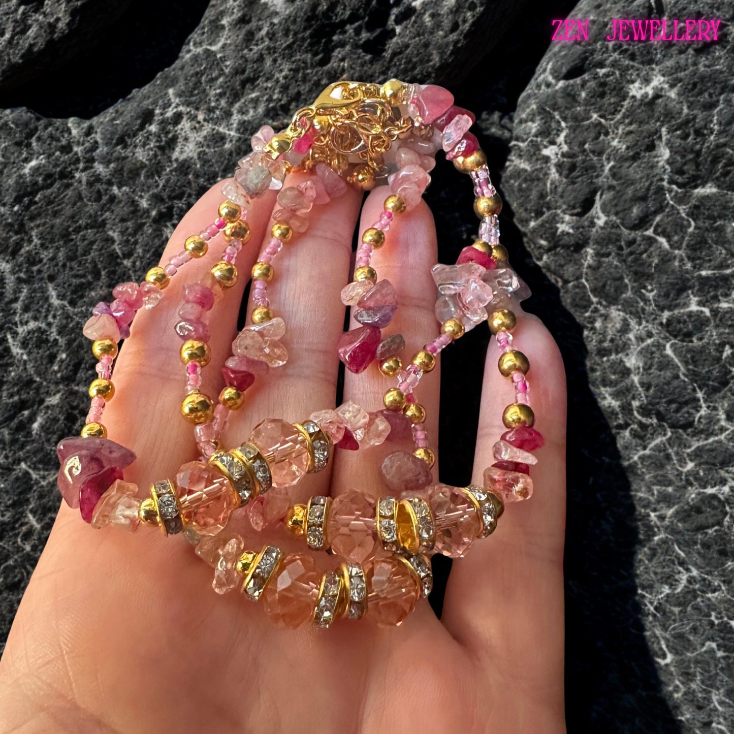 A multi-strand necklace with pink and clear gemstones, gold beads, and rhinestone detailing. The necklace has a gold clasp and is displayed on a hand. The jewellery is a mix of pink, clear, and gold colours.