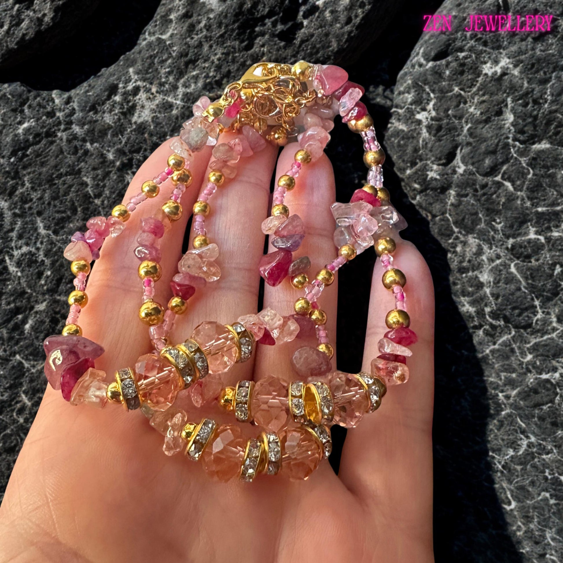 A multi-strand necklace with pink and clear gemstones, gold beads, and rhinestone detailing. The necklace has a gold clasp and is displayed on a hand. The jewellery is a mix of pink, clear, and gold colours.