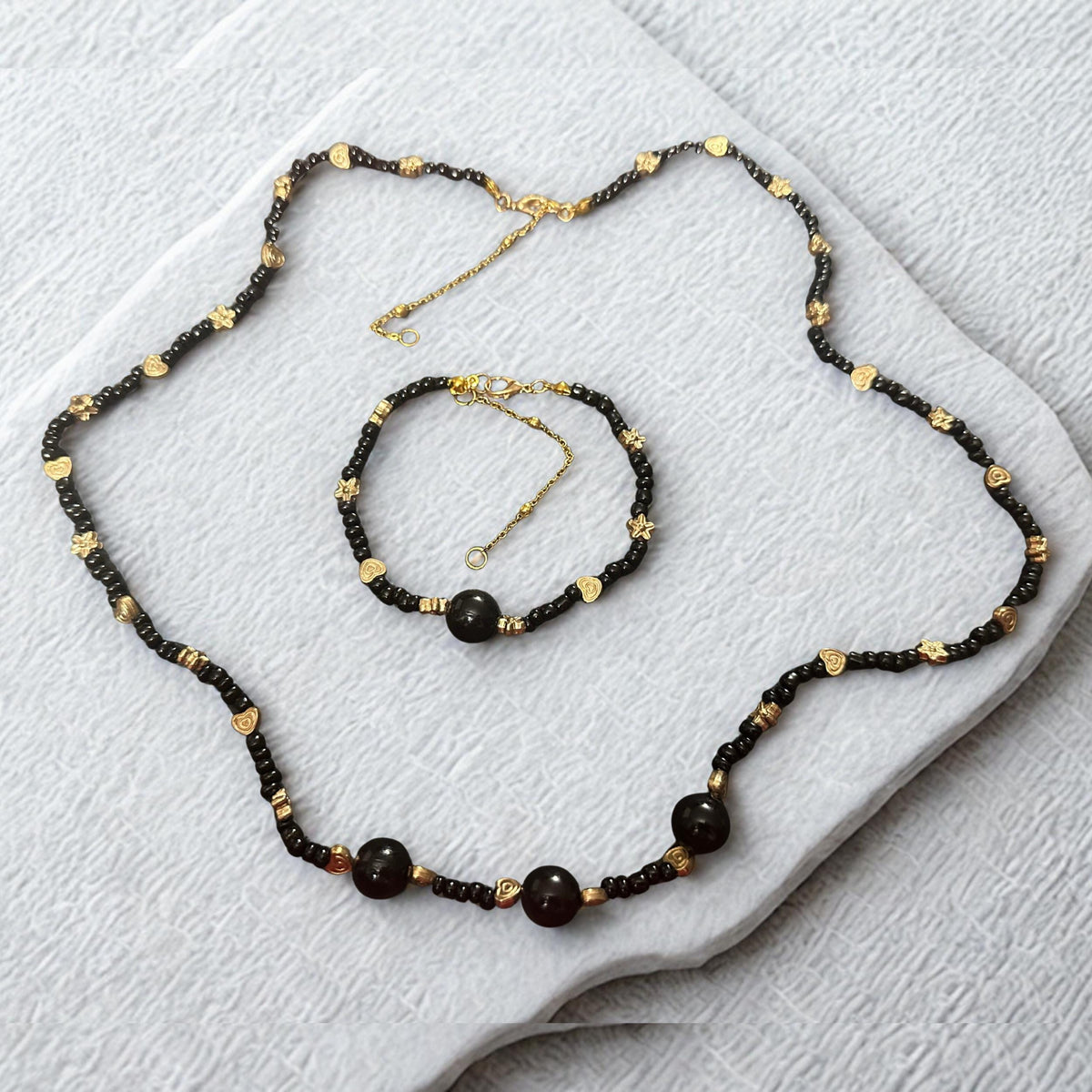 A matching bracelet accompanies the necklace and features the same premium black agate beads and gold-tone accents for a coordinated jewellery look. The bracelet includes floral spacers, decorative heart details, and a central black agate focal bead. Positioned near the necklace, it demonstrates how the two pieces complement one another perfectly. The adjustable extension chain is visible, providing insight into sizing flexibility and comfortable